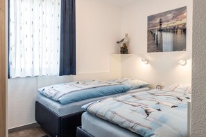 A bedroom with two beds, a window with blinds, a wall-mounted shelf with a bird figurine, and a painting of a pier.