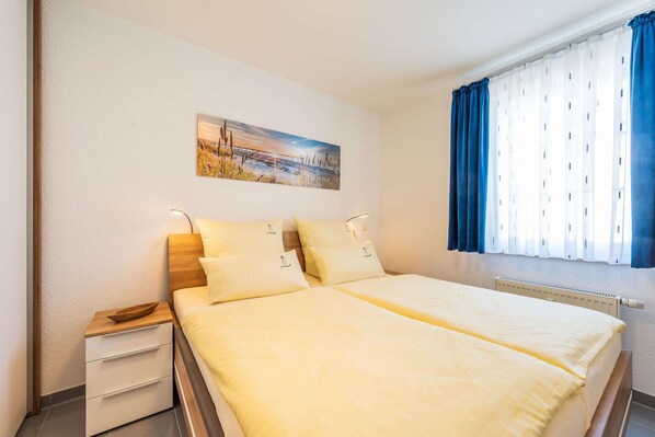 A double bed with two pillows, a bedside table, a wall-mounted painting, and a window with curtains.