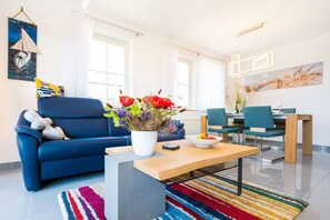 A modern living room with a blue sofa, a coffee table with a vase of flowers, a dining area with a table and chairs, and a large window with curtains.