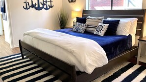 Ein Schlafzimmer mit einem Bett, einem Streifenmuster-Teppich, einer Wanddekoration in Ankerform und einem Fenster mit Blick auf ein Ziegelgebäude.