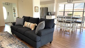 Ein modernes Wohnzimmer mit einem dunkelgrauen Sofa, einem gemusterten Teppich und einem Essbereich mit weißen Stühlen und einem Tisch.