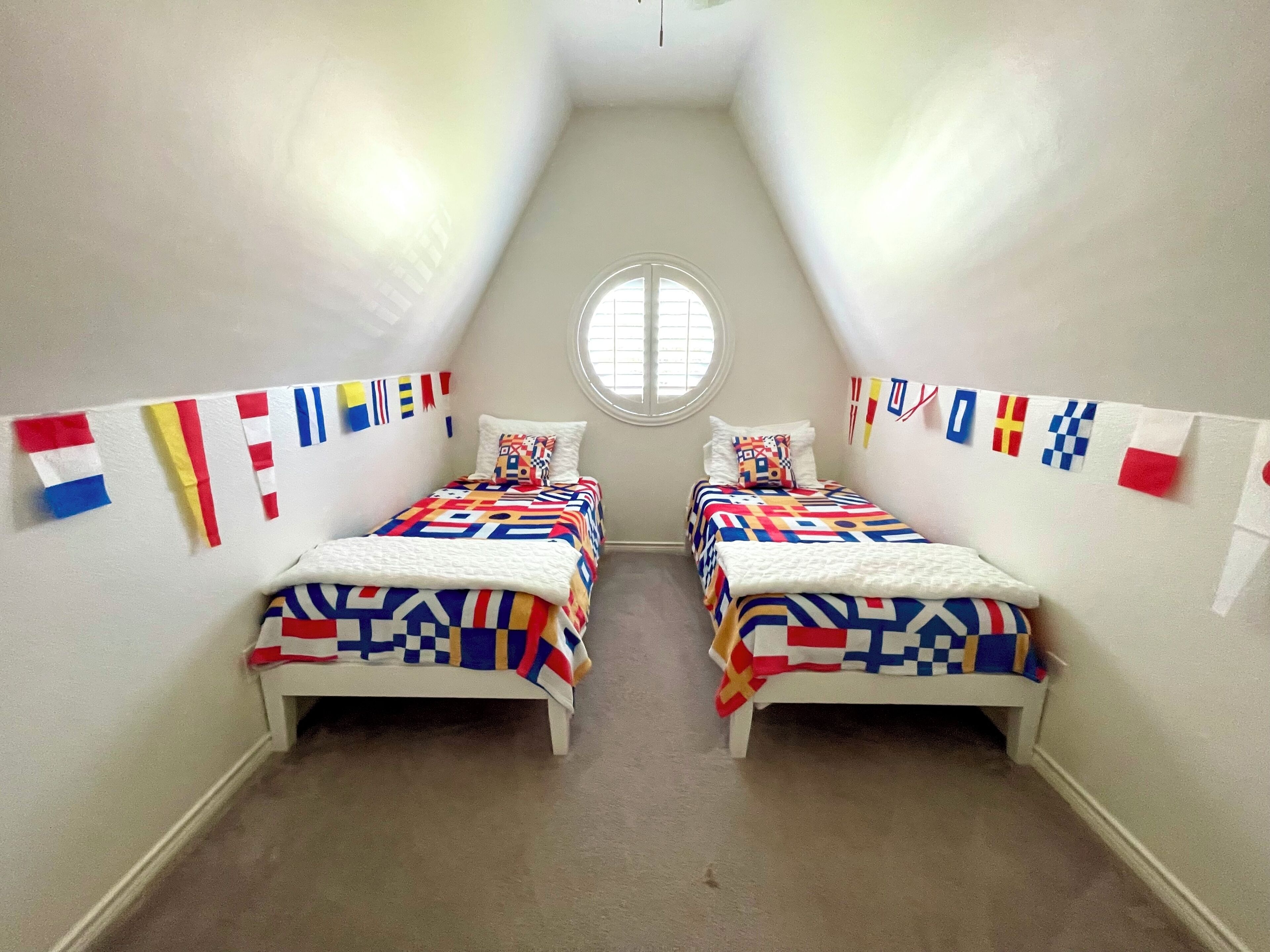 A small, narrow room with two beds, each covered with patterned bedding. There are multiple flags hanging on the walls, and a window at the far end.