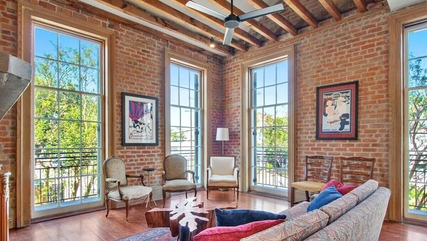 A spacious living room with large windows, a ceiling fan, and a brick wall. - Fab 1838 Garden District 5BR w/Parking & Balcony (New Orleans)