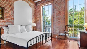 A bedroom with a brick wall, a large window, a bed with white bedding, and a wooden floor. - Fab 1838 Garden District 5BR w/Parking & Balcony (New Orleans)