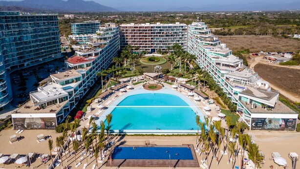 A large hotel with multiple buildings, a central swimming pool, and a beach area with sun loungers and umbrellas.