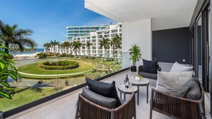 A balcony with wicker furniture, a small table, and a view of a hotel complex and a landscaped area with palm trees.