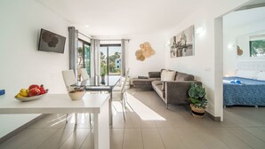 A modern living room with a glass dining table, a sofa, and a bed with a blue cover. There are plants and artwork on the walls. - Oratge Beach Views by Interhome (Port Pollença)