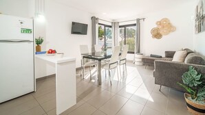 A modern kitchen with a refrigerator, a dining area with a glass table and chairs, a gray sofa, and a TV mounted on the wall. - Oratge Beach Views by Interhome (Port Pollença)
