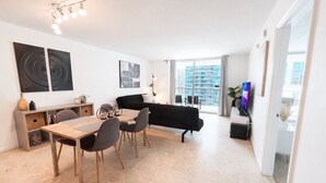 A modern living room with a dining area, a black sofa, a TV, and a large window with a city view. - 4 bed boho condo with amazing view *Comfortable, stylish and clean* (Miami)