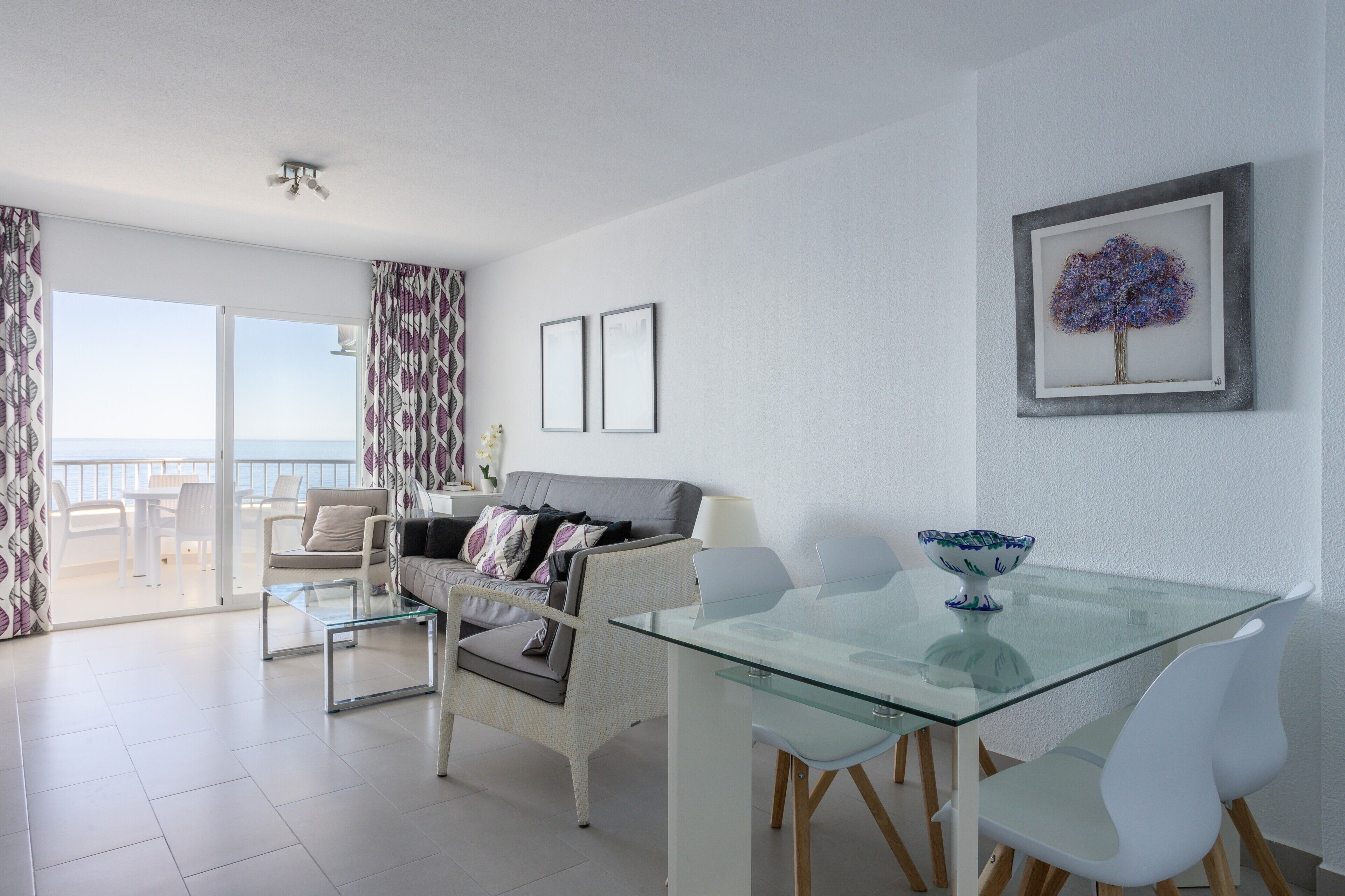 Un salon moderne avec une table à manger en verre, des chaises blanches, un canapé gris et un balcon avec vue.