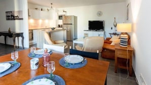 Un comedor con una mesa de madera dispuesta para cuatro, una cocina al fondo, una sala de estar con sofá y televisor, y una lámpara sobre una mesita auxiliar.