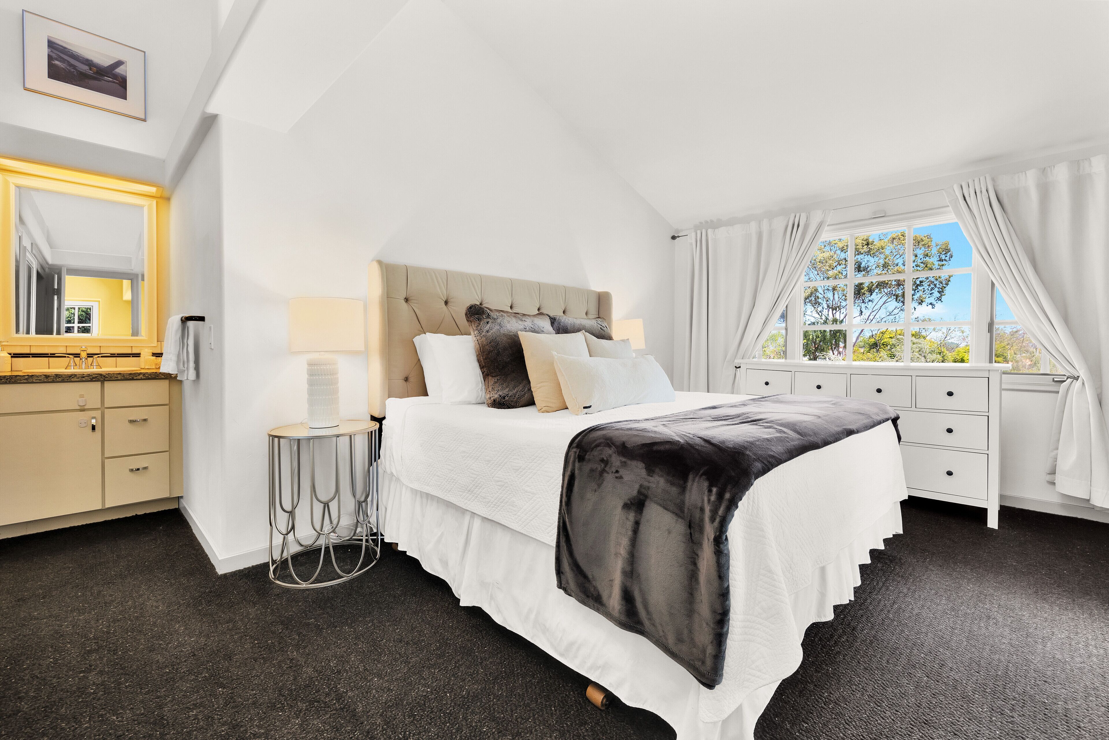 A bedroom with a large bed, a bedside table, a lamp, a dresser, and a window with curtains.