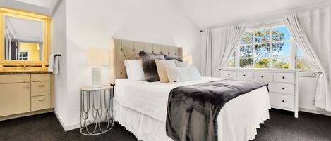A bedroom with a large bed, a bedside table, a lamp, a dresser, and a window with curtains.