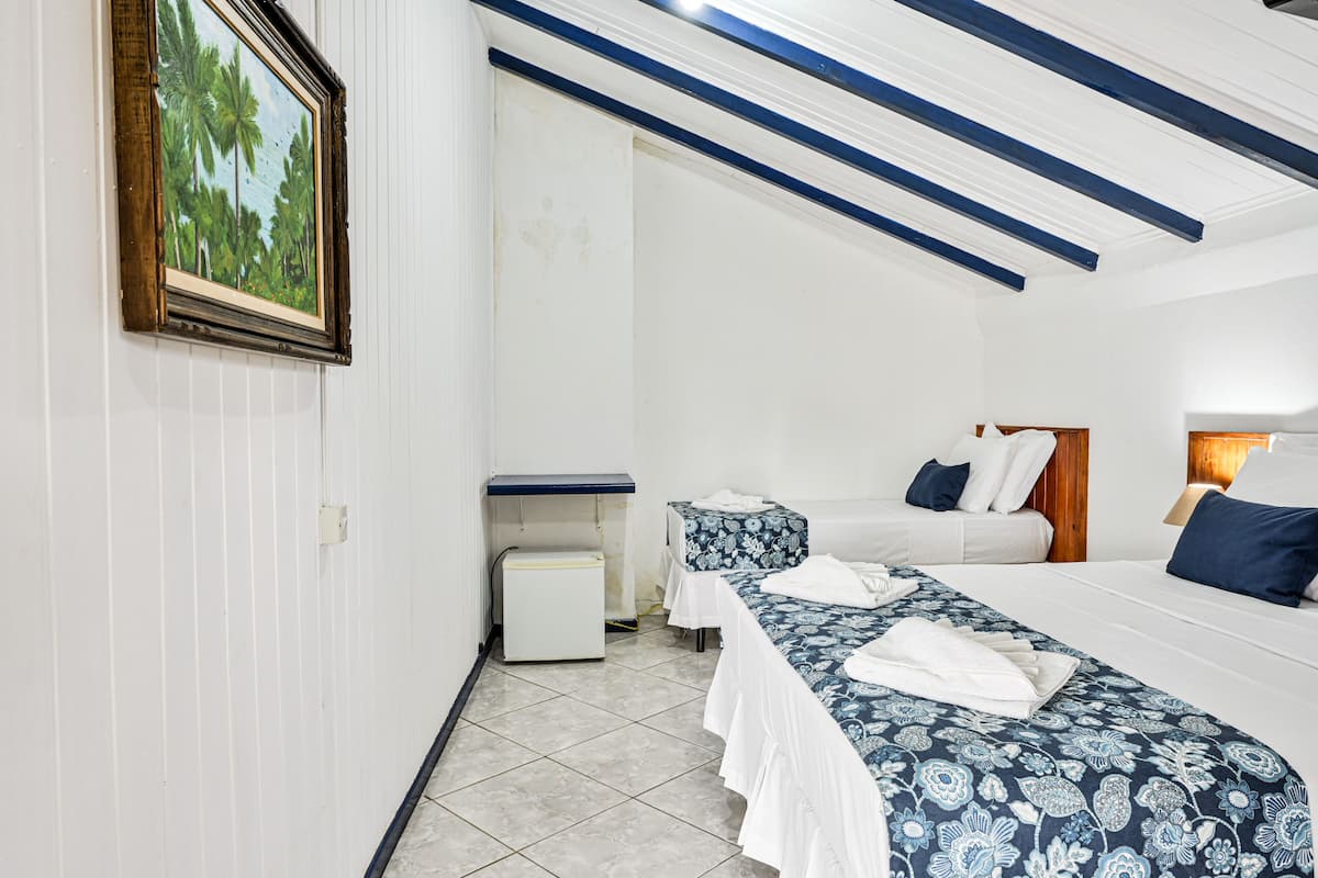 A hotel room with two beds, a small white cabinet, a painting of palm trees, and a ceiling with blue beams.
