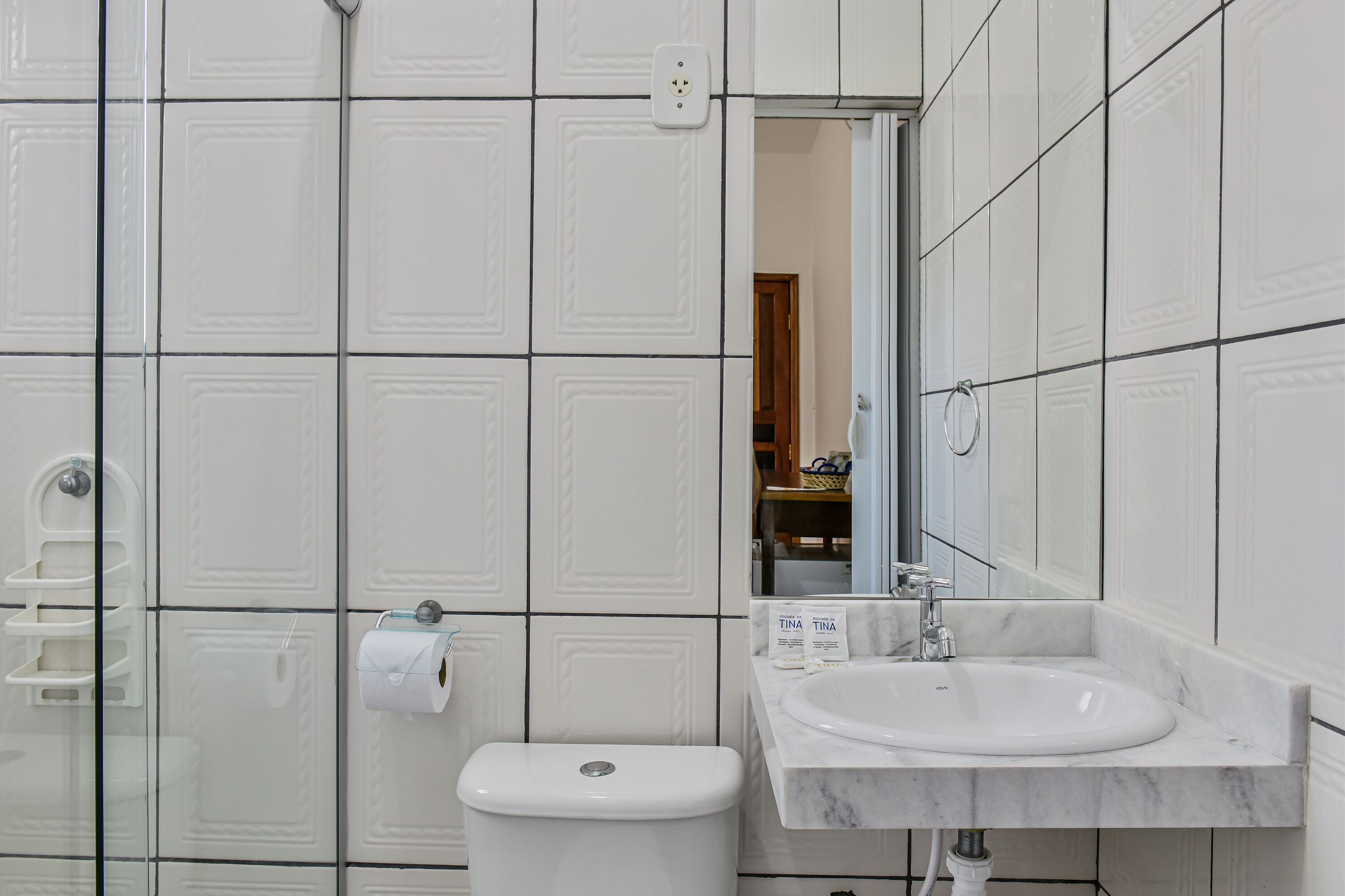 Un baño con paredes de azulejos blancos, un inodoro blanco, un lavamanos con espejo y un perchero para toallas.