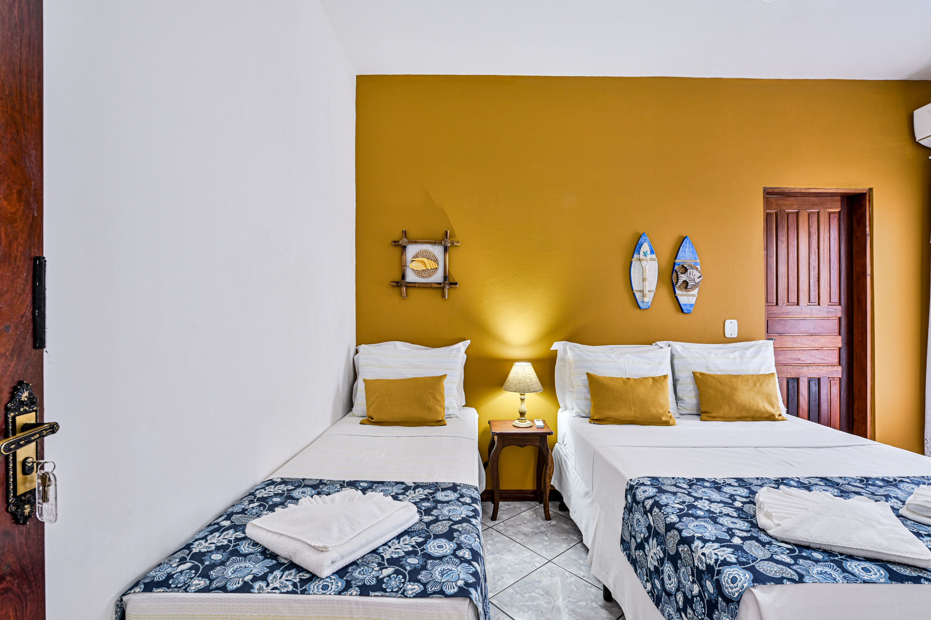 Une chambre d’hôtel avec deux lits, une porte en bois, une petite table avec une lampe et deux planches de surf accrochées au mur.