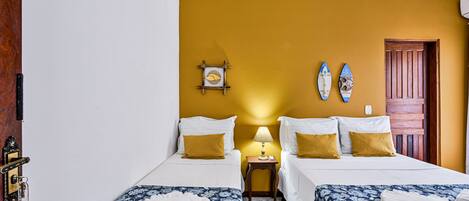 Habitación de hotel con dos camas, una puerta de madera, una mesita con lámpara y dos tablas de surf colgadas en la pared.