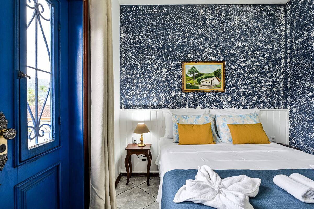 A bedroom with a blue door, a bed with white and yellow bedding, a nightstand with a lamp, and a wall with a framed picture.
