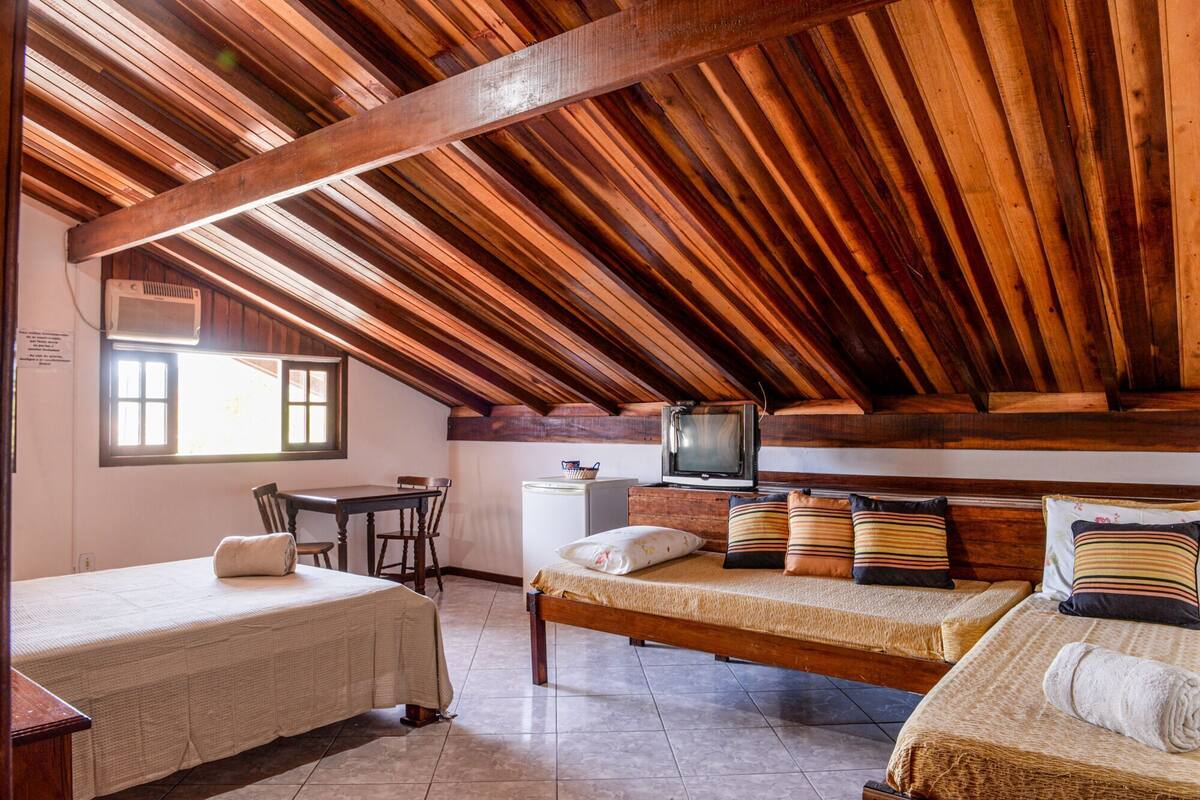A room with a sloped ceiling, wooden beams, a bed with multiple pillows, a small desk, and a television.