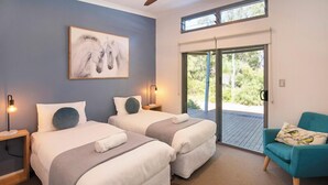 Habitación de hotel con dos camas, un ventanal amplio, una puerta corrediza de vidrio, un sillón azul verdoso y una obra de arte colgada en la pared.
