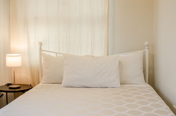 A neatly made bed with a white headboard and pillows, a bedside table with a lamp, and a curtain in the background.