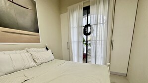 A bedroom with a bed, white curtains, a door with a wreath, and a view of the outside.