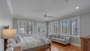 A bedroom with a bed, a nightstand, a lamp, a ceiling fan, a window with blinds, and a sofa. - Luxury, Oceanview Beach Home w/ Private Pool & Outdoor Kitchen, Hot Tub, Rooftop Deck, & Elevator! (Isle Of Palms)