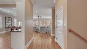 A spacious hallway with hardwood floors, a ceiling fan, and a door leading to a living area with a gray sofa and a dining area with a table and chairs. - Luxury, Oceanview Beach Home w/ Private Pool & Outdoor Kitchen, Hot Tub, Rooftop Deck, & Elevator! (Isle Of Palms)