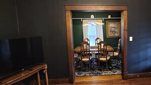 Une pièce avec un téléviseur posé sur un meuble en bois, une table à manger avec des chaises et un mur de couleur vert foncé.