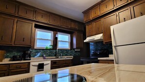 Une cuisine avec des meubles en bois, un réfrigérateur blanc, une cuisinière et une fenêtre avec des stores.