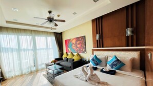 Habitación de hotel con cama, sofá, mesa de centro, ventilador de techo y un gran ventanal con cortinas.