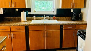 A kitchen with wooden cabinets, a granite countertop, a sink, a window with a leaf-patterned valance, and a coffee maker. - Charming home with lake views (Boydton)