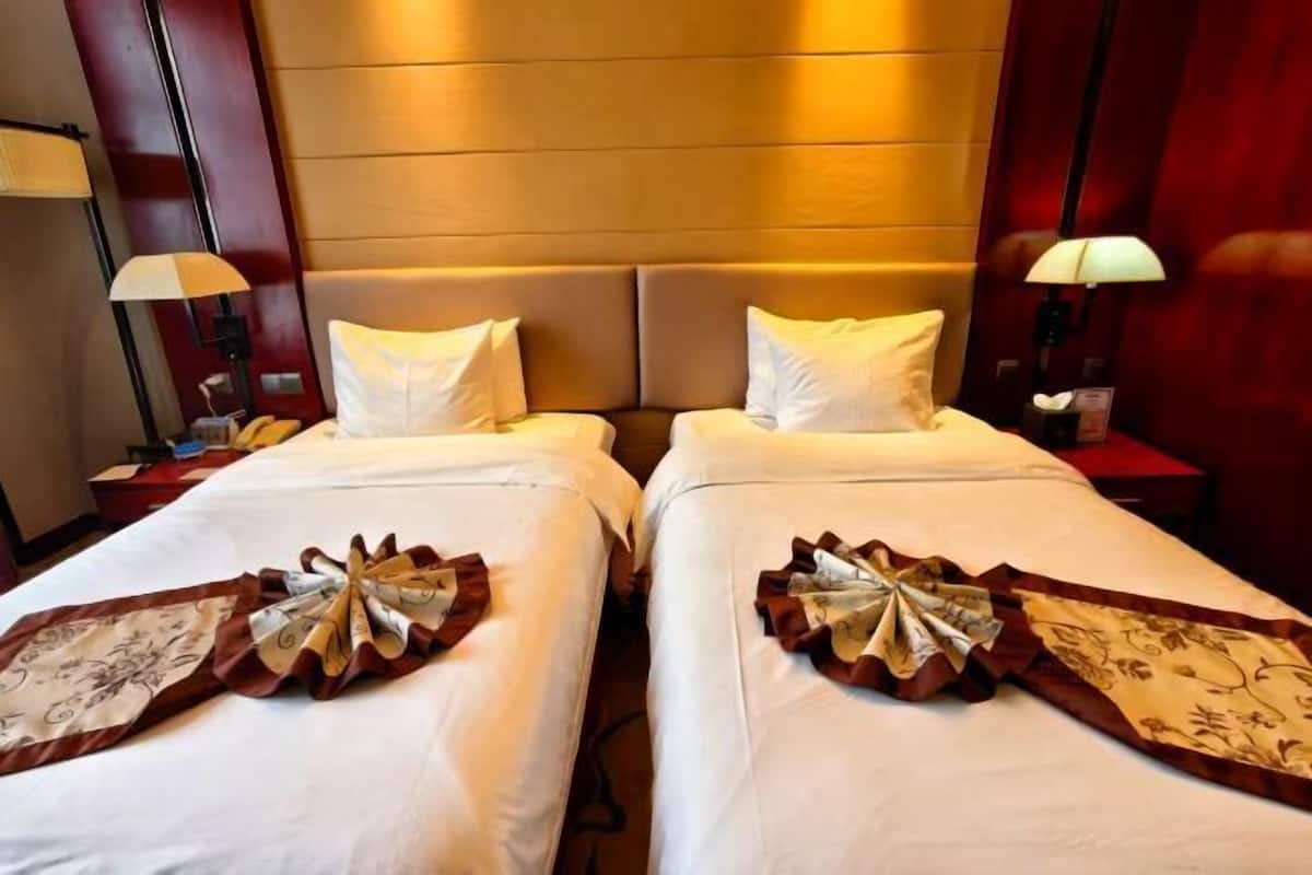 Quarto de hotel com duas camas, cada uma adornada com um laço decorativo na colcha, uma mesa de cabeceira com abajur e cabeceira fixa na parede.