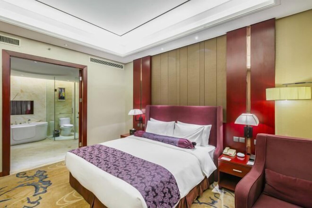A hotel room with a large bed, a bedside table with a lamp, a chair, and a bathroom visible through an open door.