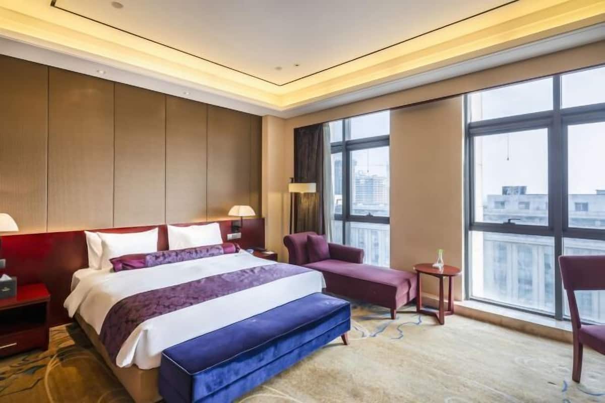 Quarto de hotel com uma cama grande, uma escrivaninha, uma cadeira, uma mesinha e vista da cidade através de amplas janelas.