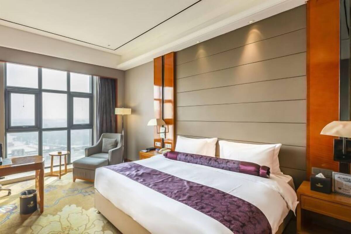 Quarto de hotel com uma cama grande, uma escrivaninha, uma cadeira, uma mesa de cabeceira e vista para a cidade através de amplas janelas.