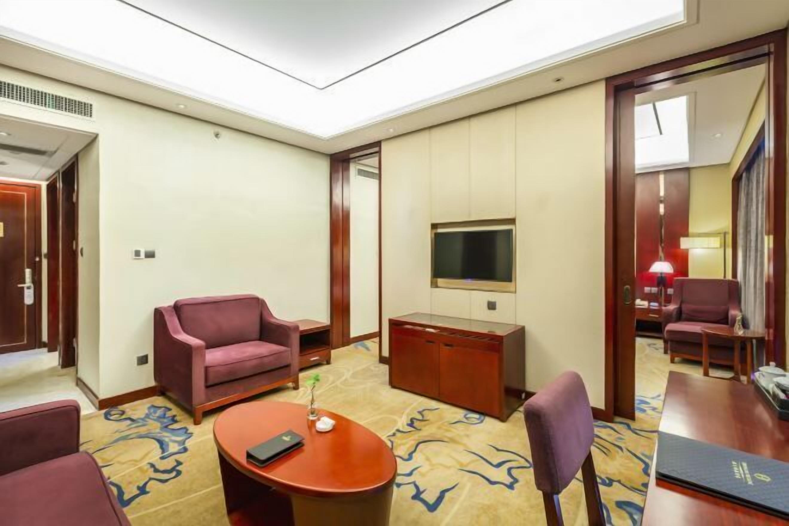 A hotel room with a flat-screen TV mounted on the wall, a wooden desk, a comfortable armchair, a sofa, and a small table with a remote control.