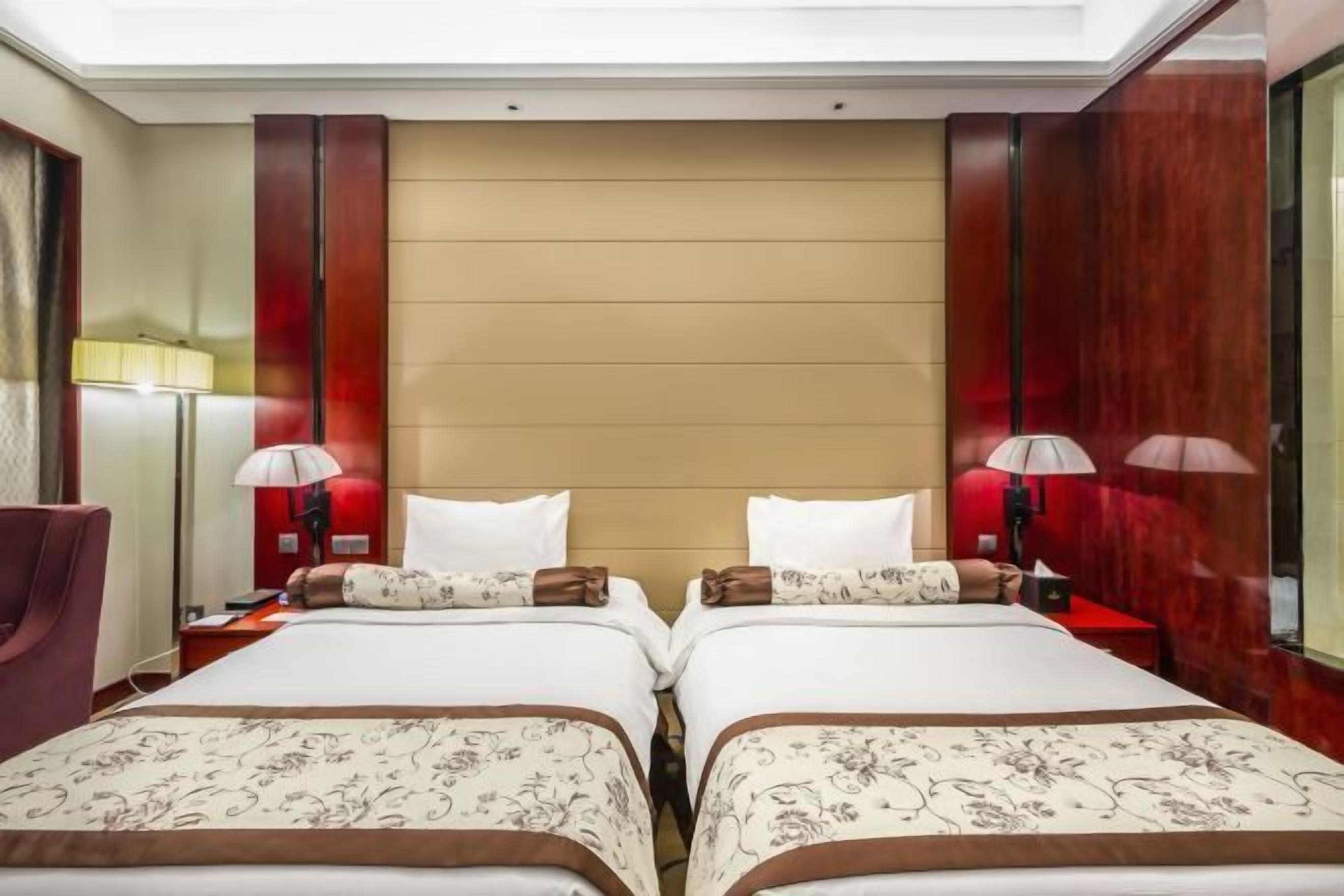 A hotel room with two beds, each with floral-patterned bedding and matching pillows. There are bedside lamps and a red-framed mirror on the wall.