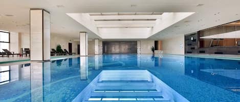 An indoor swimming pool with a clear water surface, surrounded by a modern architectural design with large windows and a high ceiling.