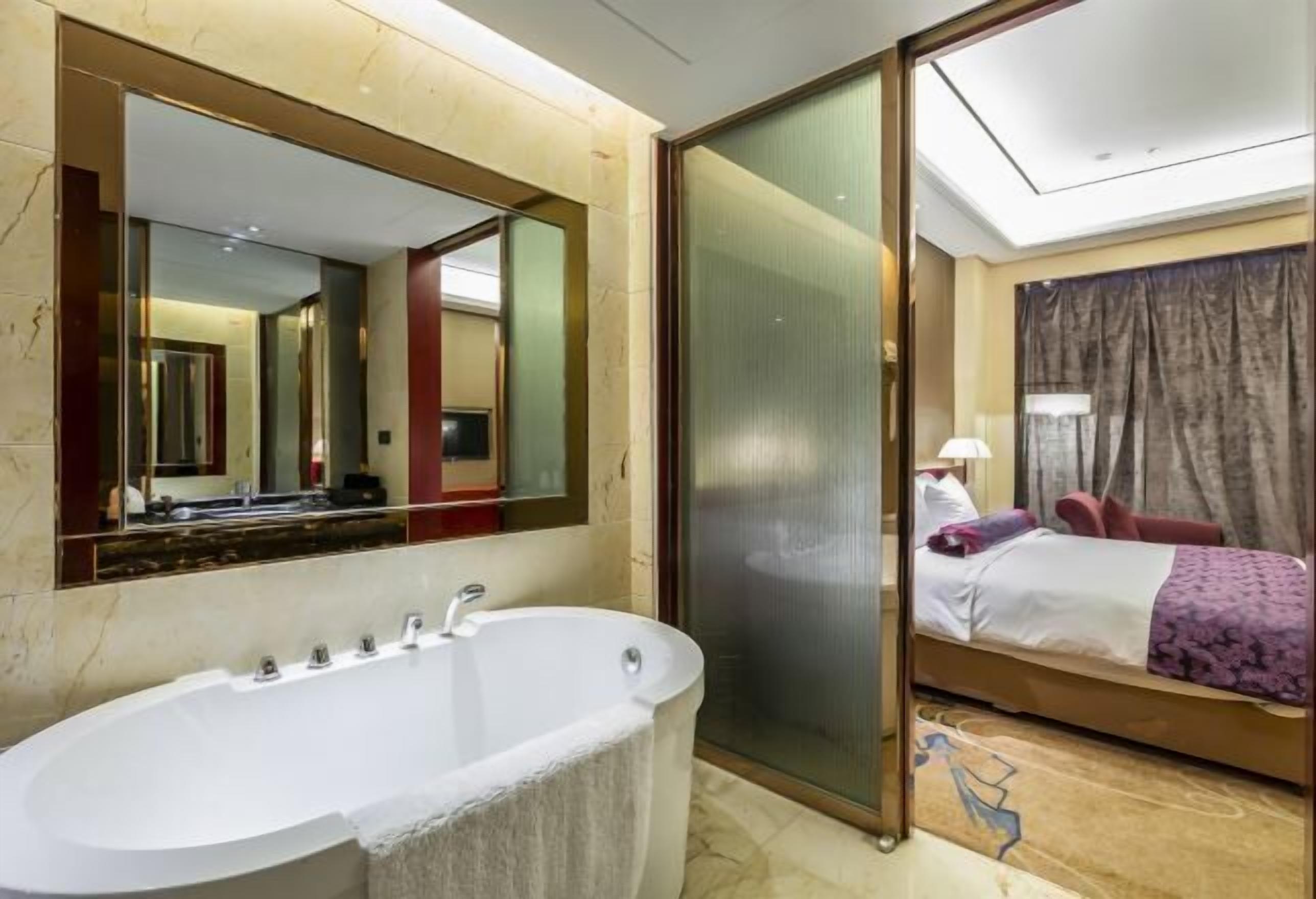 A hotel room with a large bathtub, a bed with a purple blanket, a large mirror, and a sliding door.
