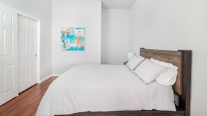 Une chambre avec un lit en bois, une literie blanche et un tableau abstrait aux couleurs vives accroché au mur.