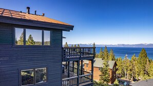 Un bâtiment moderne à plusieurs étages, doté de grandes fenêtres, d’une terrasse en bois et offrant une vue sur un lac et des montagnes.