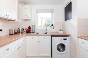 A compact kitchen with a washing machine, a toaster, a kettle, and a window with a view of the outside.