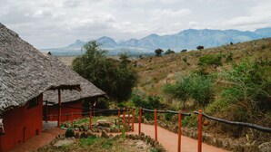 A rustic thatched-roof building with a dirt path leading to it, surrounded by natural landscape and mountains in the distance. - Leopards Lair Cottages (Mtito Andei)