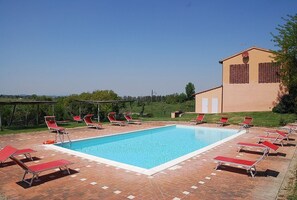 Une piscine rectangulaire entourée de chaises longues rouges, un bâtiment à la façade beige et aux volets rouges, et un ciel d’un bleu limpide.