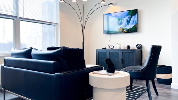 A modern living room with a black leather sofa, a round white side table, a black leather armchair, a grey cabinet with decorative items, and a large flat-screen TV mounted on the wall. - ★Black & White Themed| 1BD/1BA|Parking★ (Bound Brook)