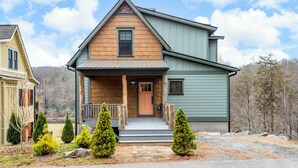 A two-story house with a wooden front and green siding, a front porch with a railing, and a small front yard with evergreen trees. - Game Room & Wraparound Decks: Cullowhee Home! (Cullowhee)