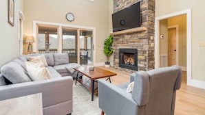 A living room with a stone fireplace, grey sofas, a wooden coffee table, and a large window. - Game Room & Wraparound Decks: Cullowhee Home! (Cullowhee)