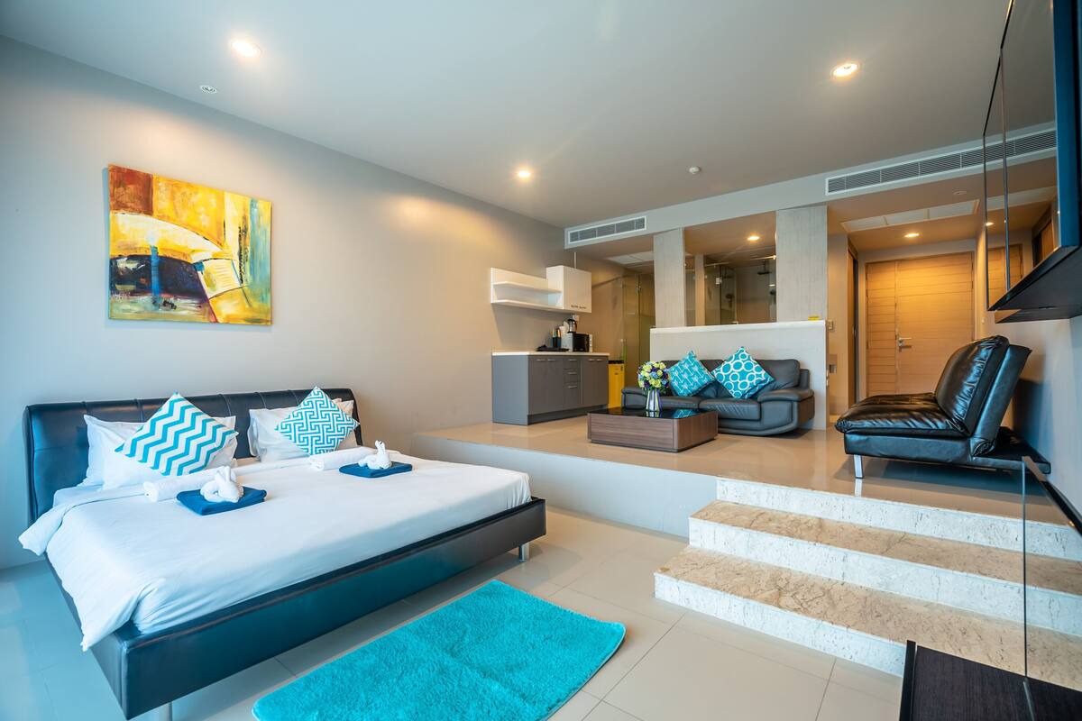 A modern hotel room with a large bed, a sofa, a coffee table, and a bathroom visible through a glass door.