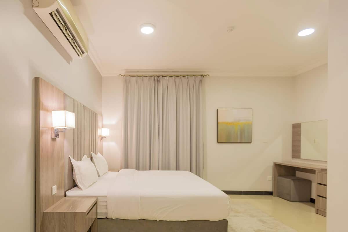 A hotel room with a bed, bedside tables, a desk, a chair, an air conditioner, a painting, and a rug.